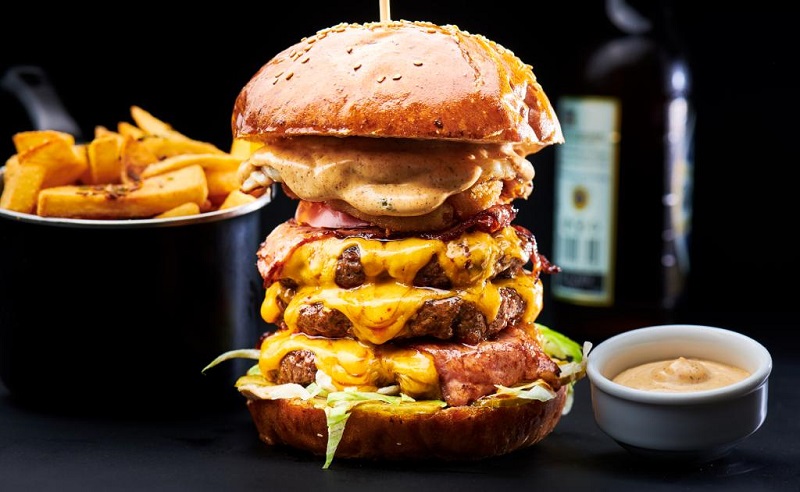 tower burger cu mai multe randuri de carne si cascaval, impreuna cu o portie de cartofi si un bol de sos, de la burger station