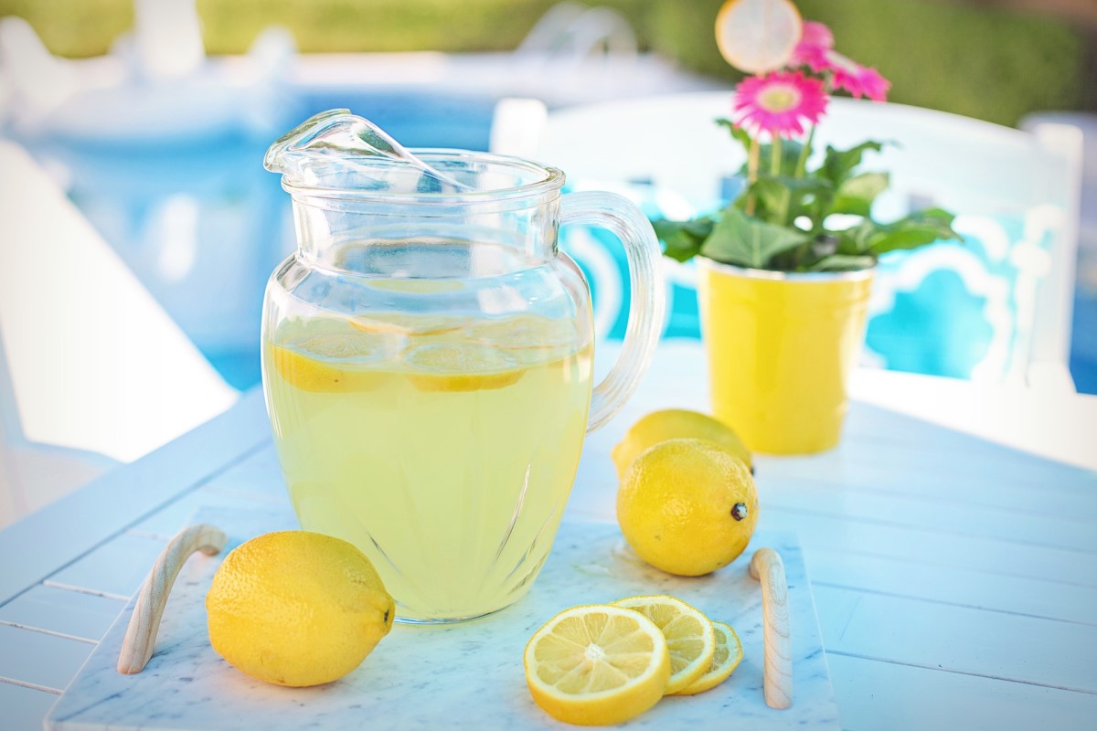 carafa cu limonada asezata pe masa, pe langa o lamaie si felii de lamaie si un ghiveci galben cu floare roz
