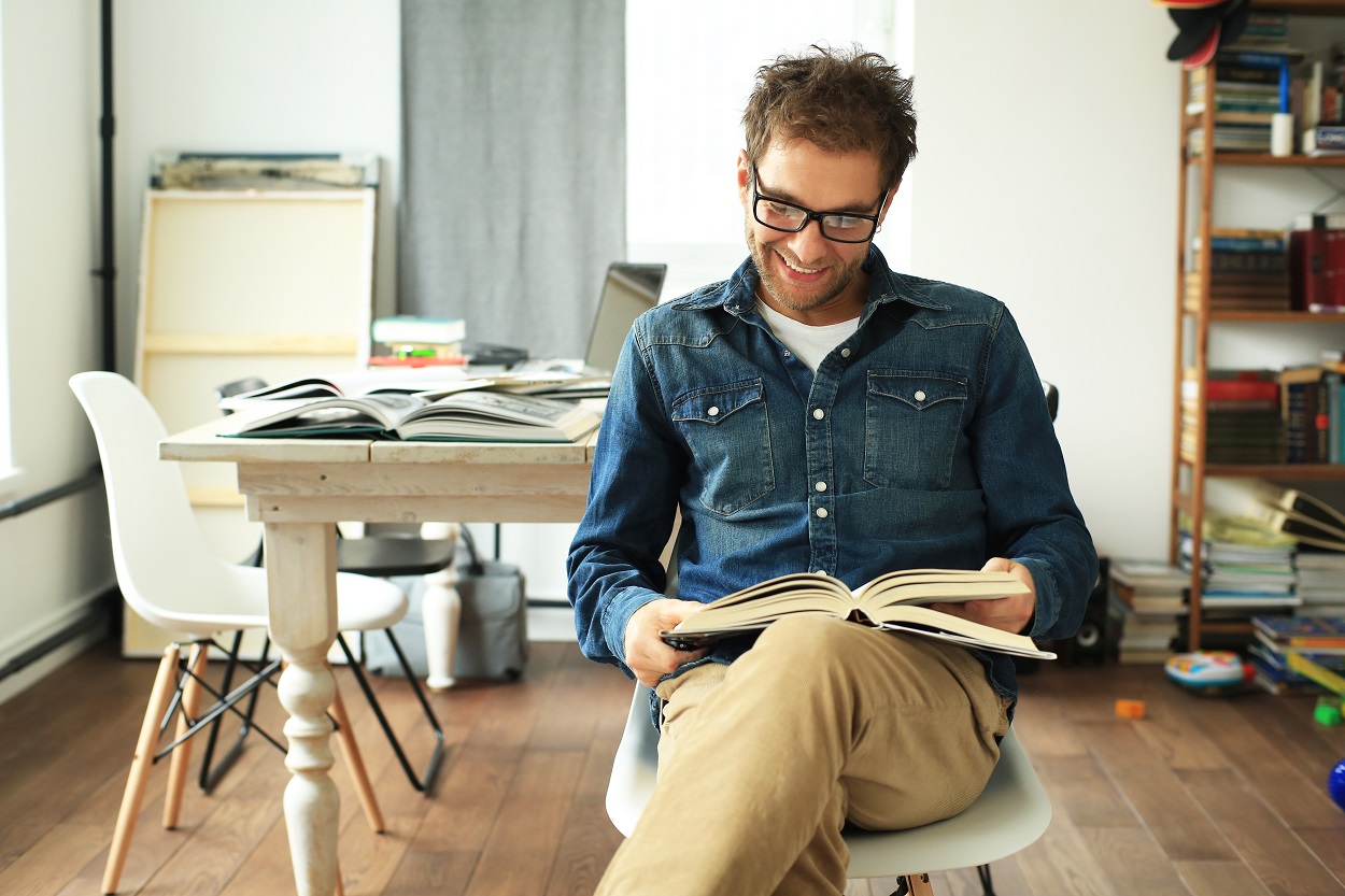 barbat tanar cu ochelari citind cărți de dezvoltare personală, așezat pe un scaun, cu mobilă în fundal, imbracat cu camasa albastra de blugi