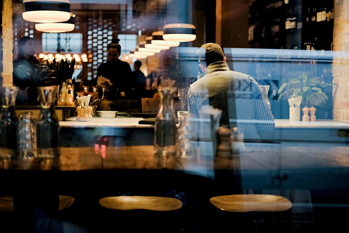 restaurant fotografiat prin vitrina, inauntrul caruia se vede un om asezat la bar si mai multe corpuri de iluminat insirate deasupra barului, situația restaurantelor în pandemie