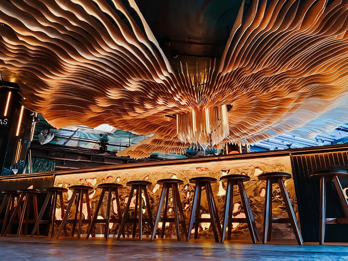 Imagine cu barul de la Naive Bucharest, cu scaune de bar insirate in fata barului luminat si un tavan plin de o sculptura din lemn asemanatoare cu un evantai