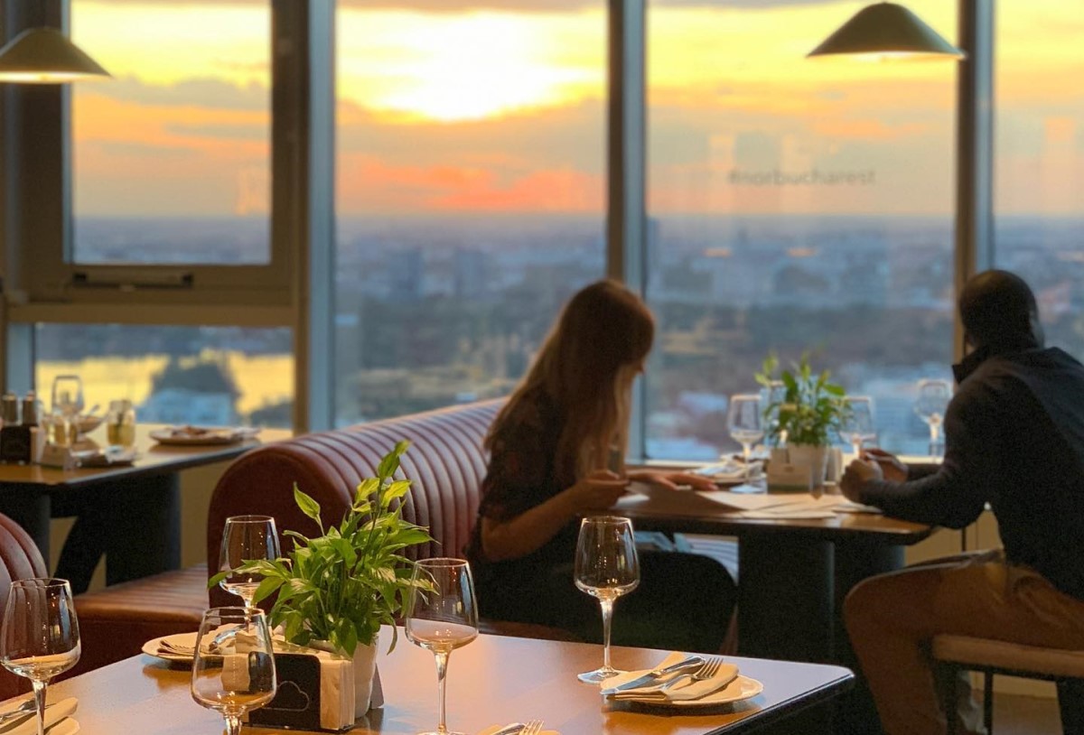 Cuplu care ia masa la restaurant in fata unor geamuri mari, la apus, cu vedere panoramica asupra orasuljui