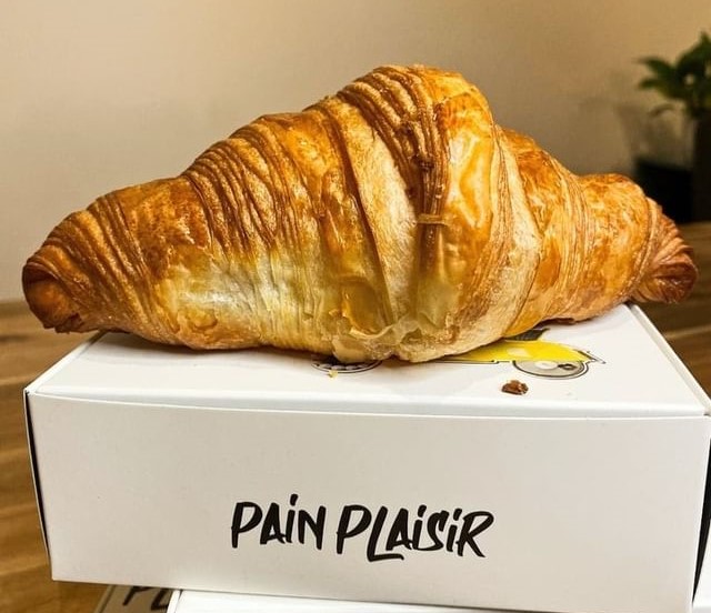 un croissant intreg asezat pe o cutie alba pe care e scris cu negru Pain Plaisir