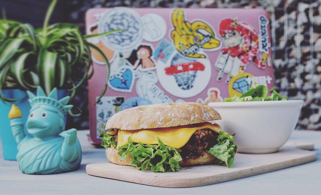 sandwich Pulled Pork, unul din burgeri de la flipflop, cu carne de vita, salata si branza cheddar, asezat pe un tocator de lemn, langa o vaza albastra, in forma de ratusca, cu un bol langa
