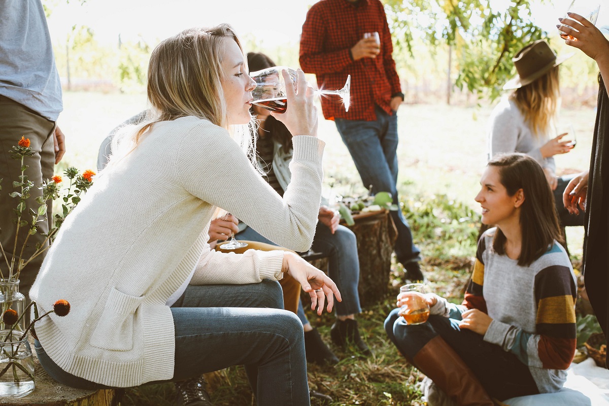 grup de tineri in natura, cu o femeie in prim plan, care bea dintr-un pahar de vin - recomandări de weekend