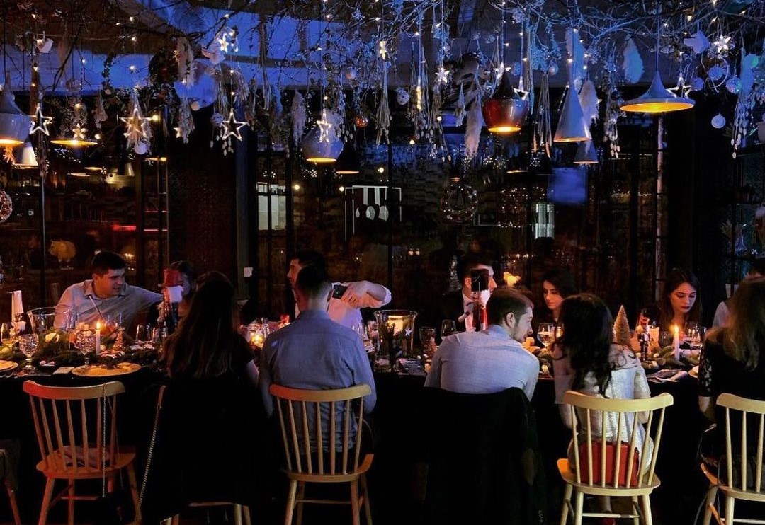 oameni asezati la o masă la restaurant cu colegii pe final de an, de Crăciun, cu lumina difuză și plante atârnate de tavan