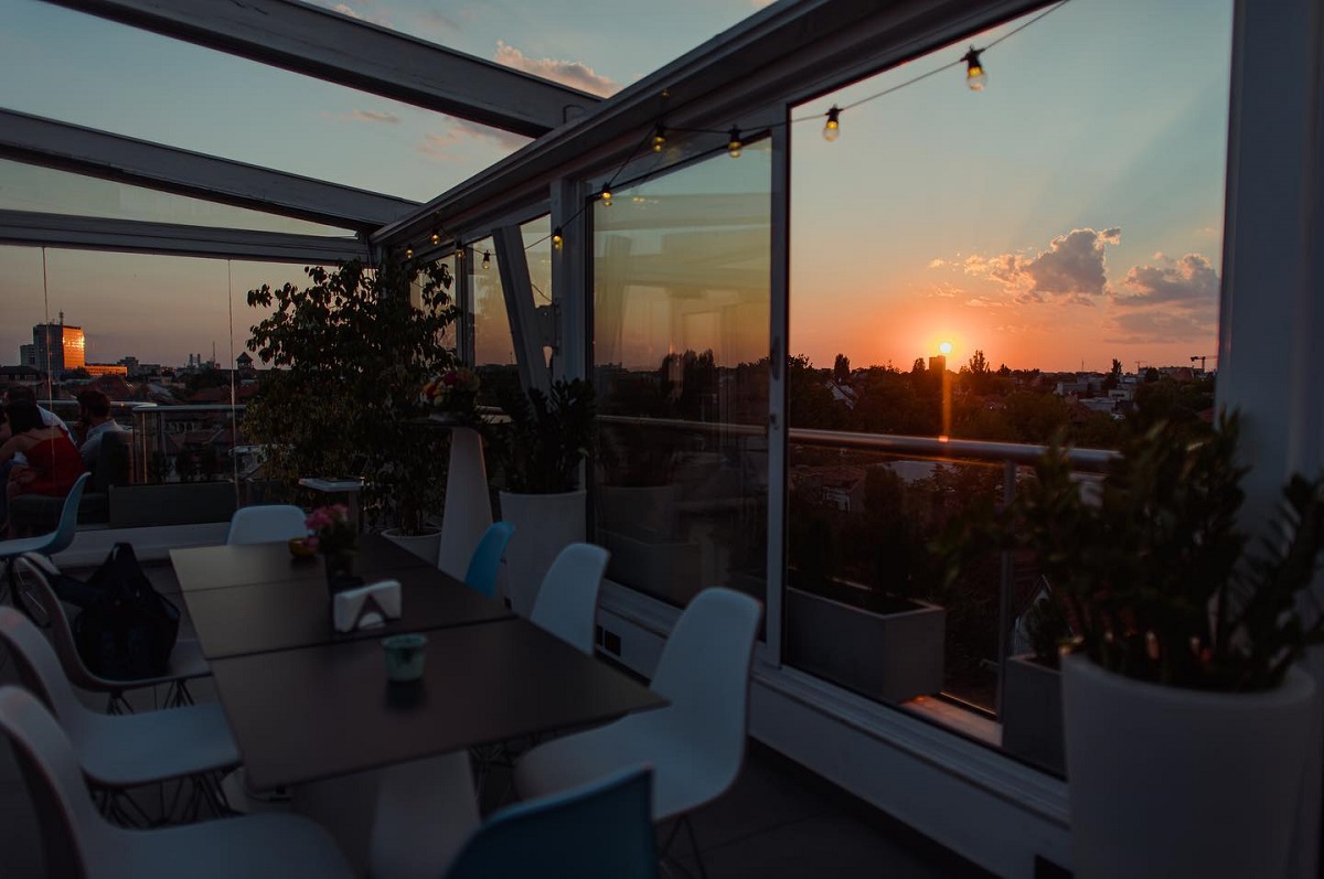 terasa sky bar dorobanti, cu o masa in prim plan si scaune albe, cu ferestre glsiante in spate, prin care se vede panorama si apusul, unul din restaurante la înălțime din București