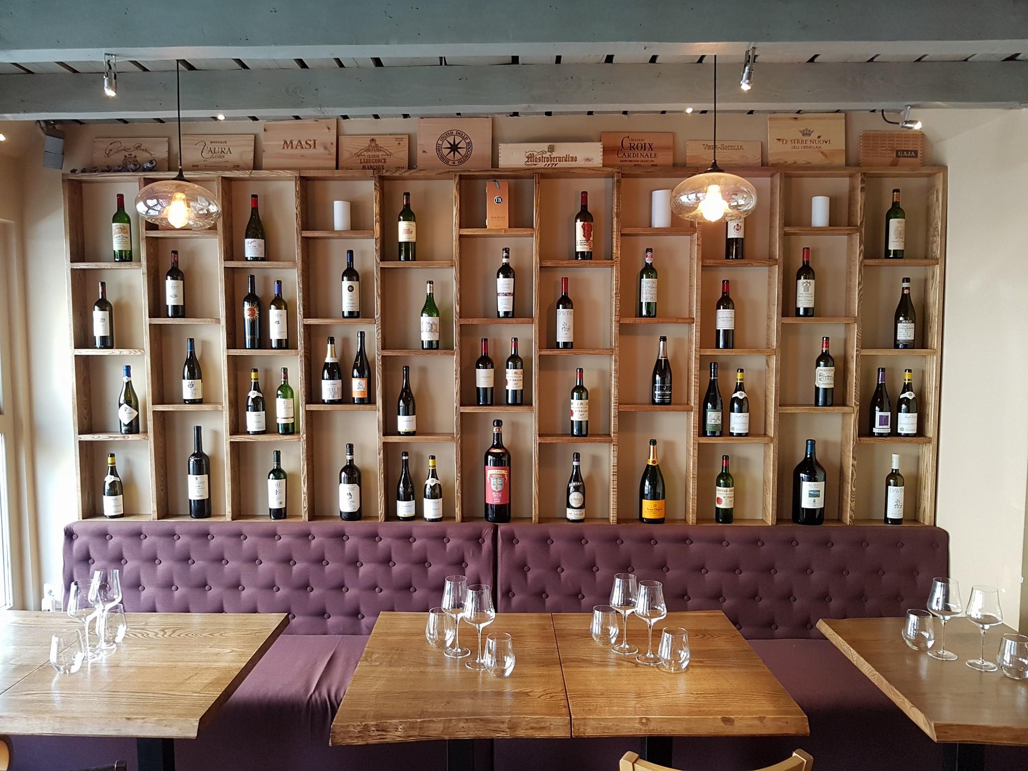 imagine din tasting room by ethic wine, cu un perete acoperit cu rafturi in care sunt expuse multe sticle de vin, in fata caruia se afla o canapea lunga, moc, capitonata, si 3 mese patrate, de lemn, pe una din ele stand asezate pahare goale de vin