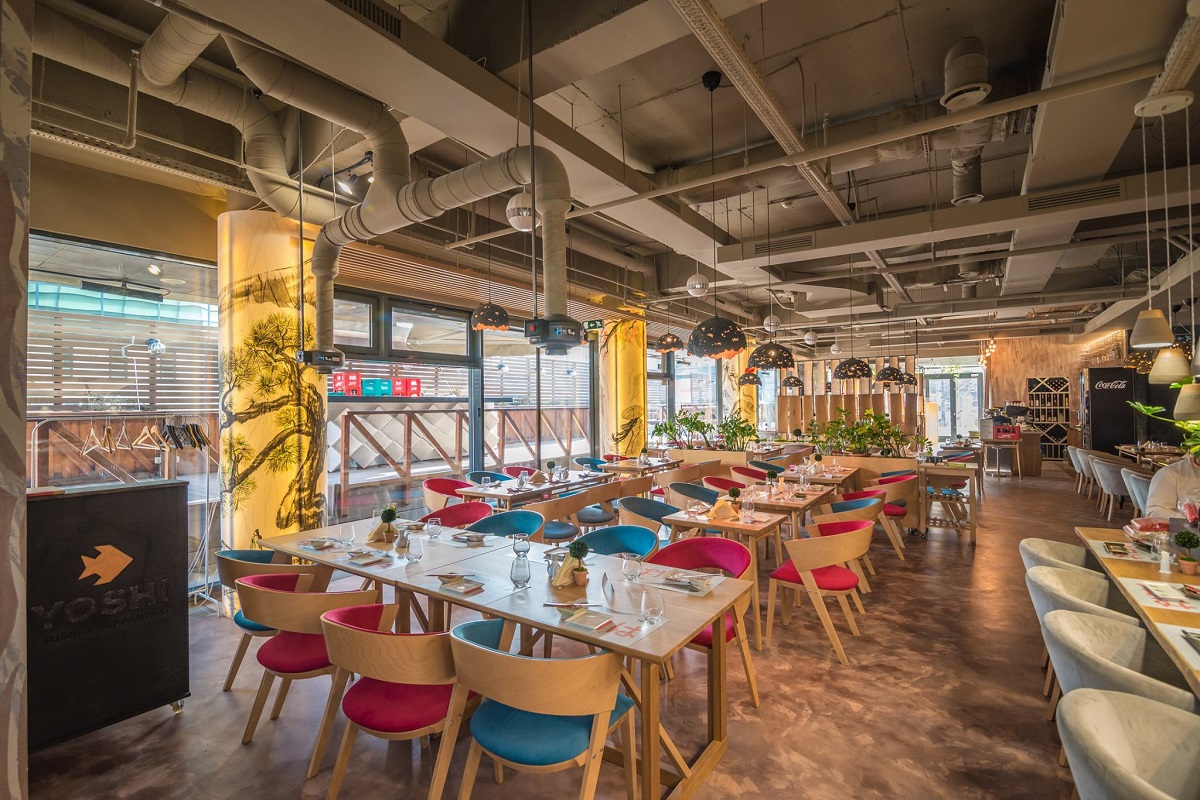 imagine de ansamblu din interiorul restaurantului Yoshi Sushi, cu mese de lemn si scaune tapitate in rosu si albastru, cu tavan industrial