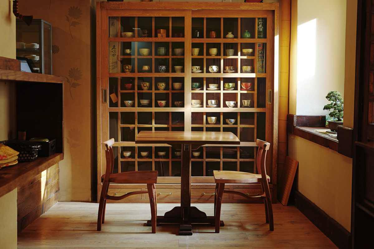 imagine din restaurant yuki japanese home dining, cu o masa cu doua scaune fata in fata, asezate in fata unui dulap cu usi de sticla, plin de vase japoneze pentru servit masa, si o fereastra in partea dreapta