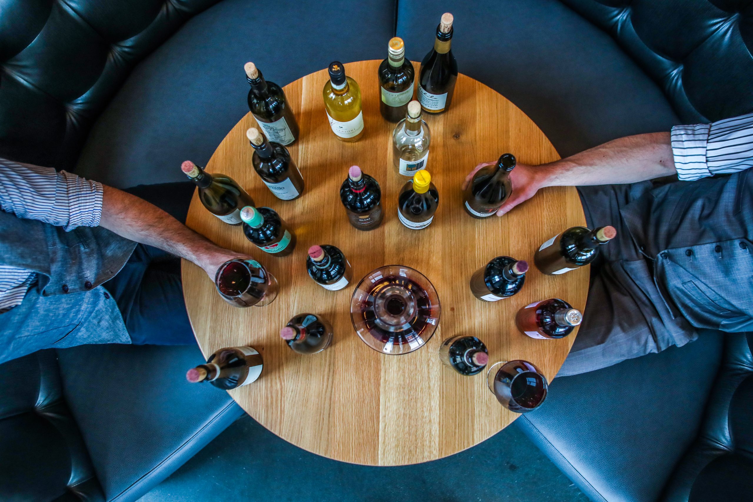 masa rotunda fotografiata de sus, pe fundal albastru, plina cu sticle de vin si doi oameni carora li se vad mainile servind un pahar de vin