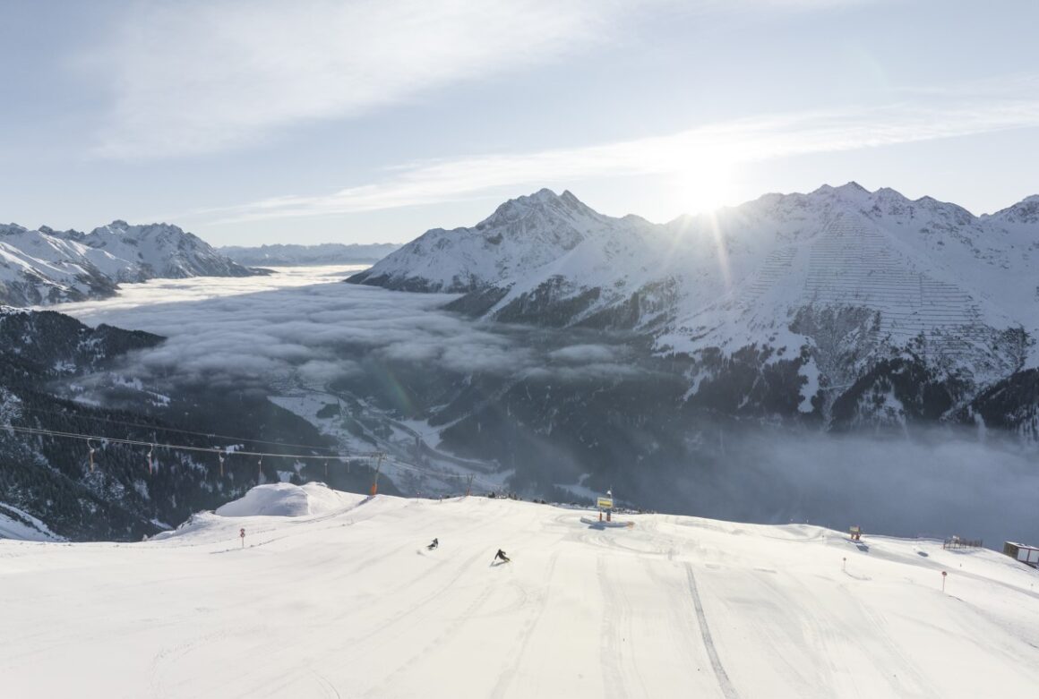 Varful muntilor din St. Anton am Arlberg, acoperiti de zapada, deasupra plafonului de nori, si cativa schiori pe partie. Una din cele mai faimoase stațiuni de ski din Europa.